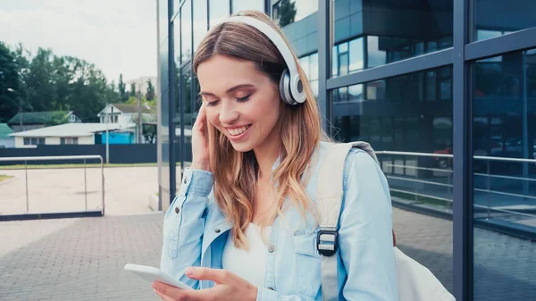 Joven mujer alegre en auriculares inalámbricos usando teléfono inteligente en la calle urbana - foto de stock