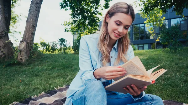 Fröhliche Studentin liest Buch, während sie auf karierter Decke im Park sitzt — Stockfoto