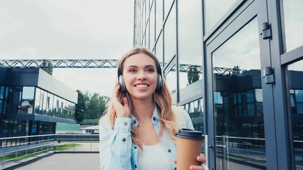Femme heureuse dans les écouteurs tenant du café pour aller tout en marchant dans la rue urbaine — Photo de stock