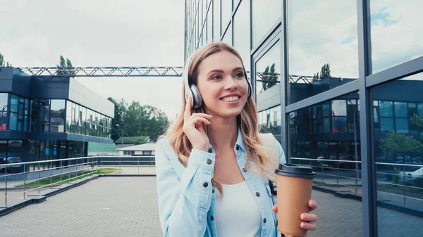 Mulher alegre em fones de ouvido sem fio segurando café para ir enquanto caminha na rua urbana — Fotografia de Stock