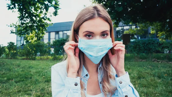 Jeune étudiant ajustant masque médical bleu dans le parc vert — Photo de stock