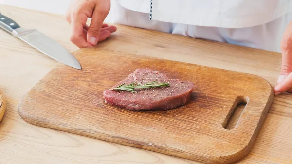 Partial view of chef standing near beef steak with rosemary branch on chopping board — Stock Photo