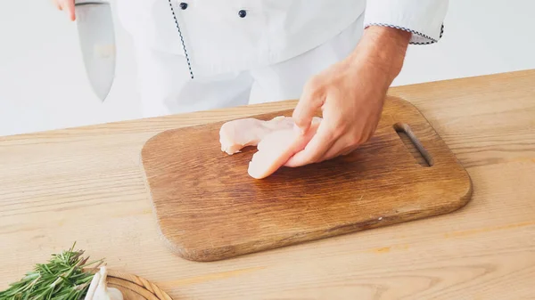 Vista recortada del chef cortando filete de pollo cerca de los ingredientes en la mesa en blanco - foto de stock
