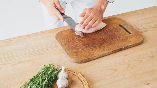 Vista parcial del hombre cortando filete de pollo cerca de los ingredientes en la mesa en blanco - foto de stock