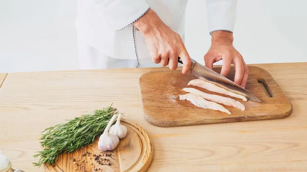 Vue partielle du chef coupant le filet de poulet sur la planche à découper sur blanc — Photo de stock