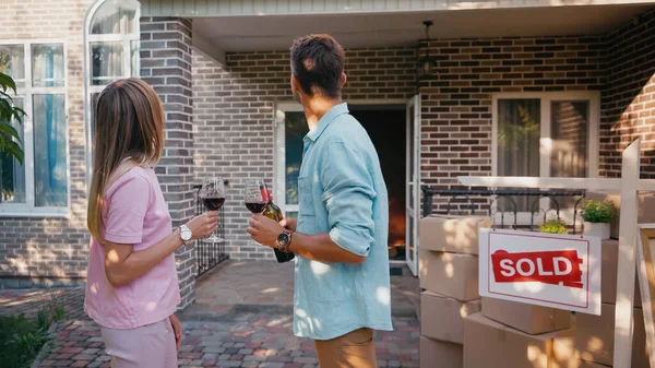 Visão traseira do casal segurando copos de vinho tinto perto da placa com letras vendidas e nova casa — Fotografia de Stock