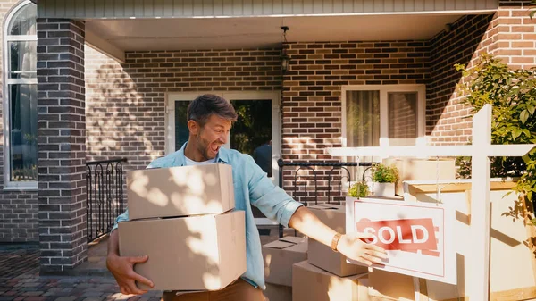Excited man holding boxes and pointing with hand at sold board near house — Stock Photo