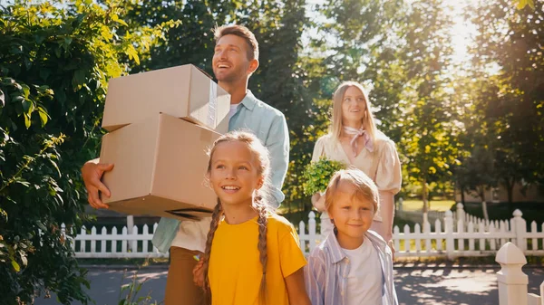 Cheerful parents holding boxes and plant while walking with happy kids, relocation concept — Stock Photo