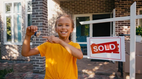 Enfant joyeux pointant vers les clés près de bord avec lettrage vendu et nouvelle maison — Photo de stock