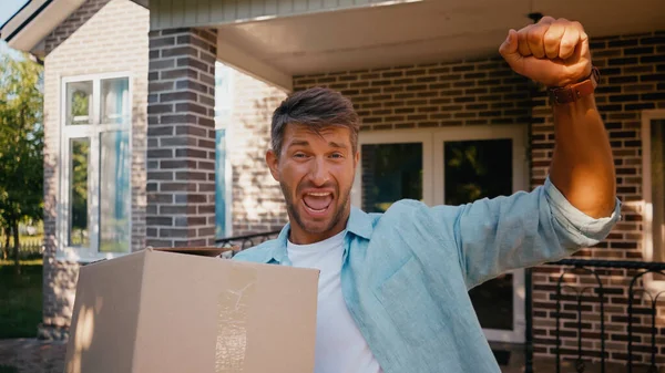 Hombre excitado sosteniendo caja de cartón y regocijo cerca de casa nueva - foto de stock