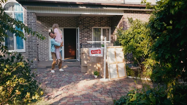 Счастливый мужчина поднимает жену рядом с коробками и новым домом — стоковое фото