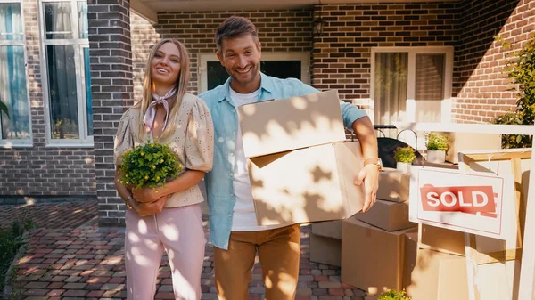 Alegre pareja con cajas y planta de pie cerca de nueva casa - foto de stock