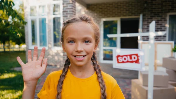 Радісна дитина дивиться на камеру і махає рукою біля розмитої дошки з проданим написом і новим будинком — стокове фото