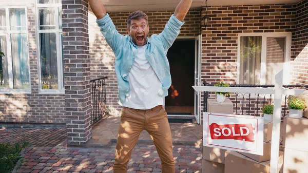 Homme excité avec les mains levées et bouche ouverte près de bord avec lettrage vendu et nouvelle maison — Photo de stock