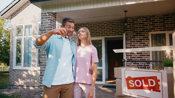 Hombre feliz sosteniendo las llaves y abrazando a la esposa cerca de la nueva casa y el tablero vendido - foto de stock