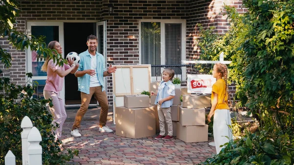 Mother throwing football to kids near husband, carton boxes and new house — Stock Photo