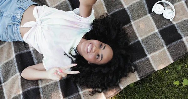Draufsicht einer glücklichen afrikanisch-amerikanischen Frau, die auf Picknickdecke liegend ein Friedenszeichen zeigt — Stockfoto