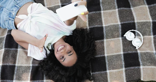 Vista superior de la alegre mujer afroamericana tomando selfie en el teléfono inteligente mientras está acostado en la manta de picnic - foto de stock