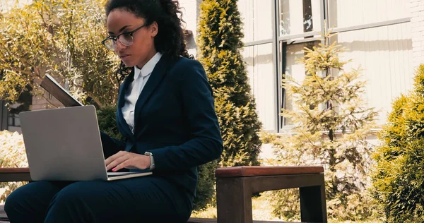 Femme d'affaires afro-américaine bouclée utilisant un ordinateur portable et tenant un carnet tout en étant assis sur le banc — Photo de stock