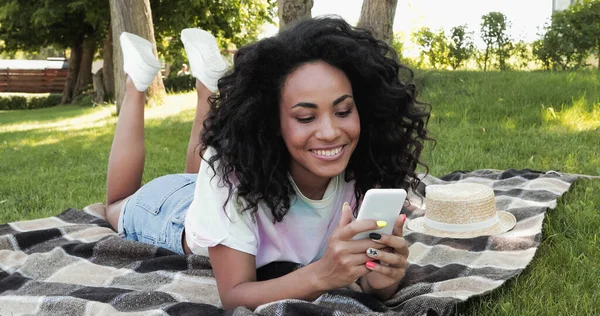 Sonriente mujer afroamericana usando un teléfono inteligente mientras está acostado en una manta a cuadros en el parque - foto de stock
