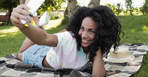 Femme afro-américaine joyeuse prenant selfie sur smartphone tout en étant allongé sur une couverture dans le parc — Photo de stock