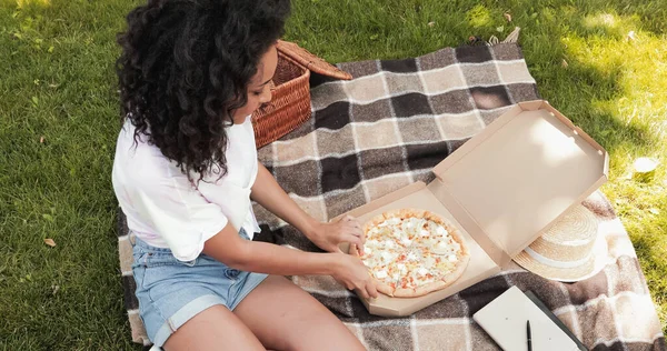 High angle view of happy african american woman reaching pizza during picnic in park — Stock Photo