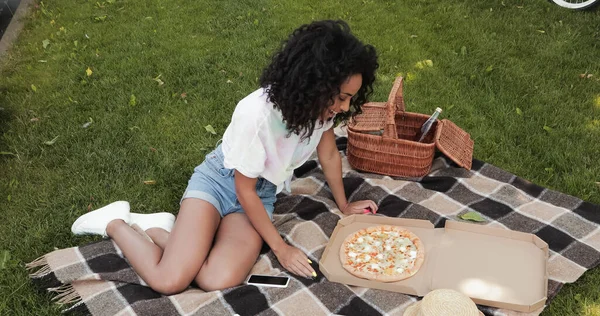 Высокий угол зрения счастливой африканской американки глядя на пиццу в коробке во время пикника в парке — стоковое фото