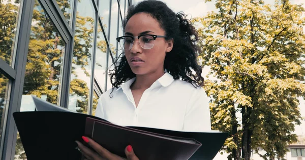 Mujer de negocios afroamericana buscando carpetas cerca del edificio exterior - foto de stock