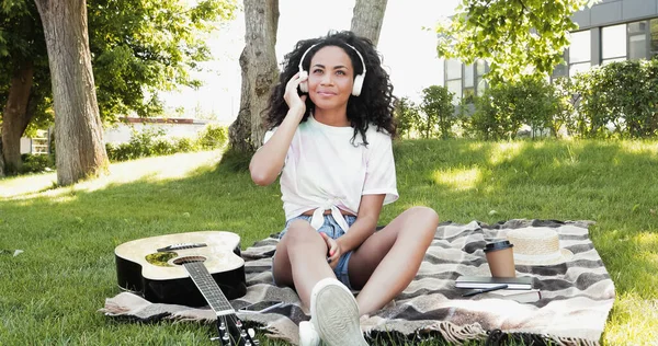 Mujer afroamericana sonriente en auriculares escuchando música durante el picnic en el parque - foto de stock