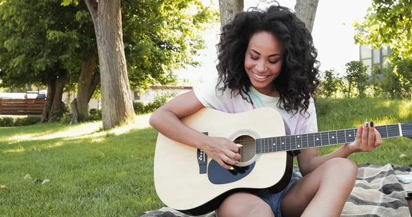 Sonriendo mujer afroamericana tocando la guitarra acústica en el parque - foto de stock