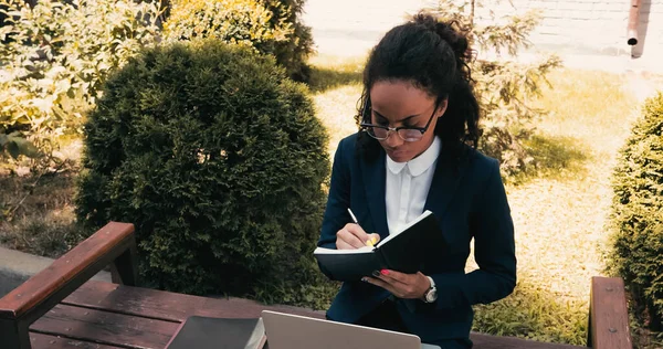 Femme d'affaires afro-américaine écrit dans un cahier et assis sur un banc avec ordinateur portable — Photo de stock