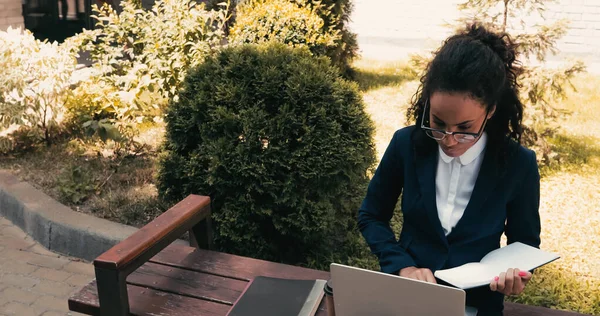 Afroamerikanische Geschäftsfrau mit Brille hält Notizbuch in der Hand und sitzt auf Bank mit Laptop — Stockfoto