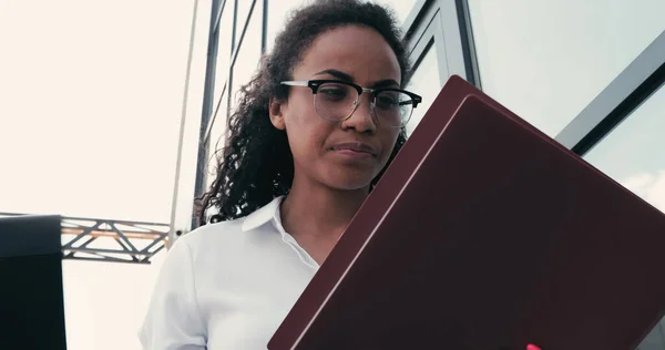 Low angle view of young african american businesswoman in eyeglasses looking at folders near building — Stock Photo