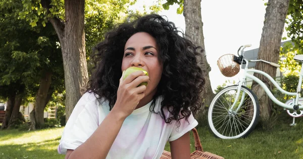 Young african american woman eating fresh apple near bicycle in park — Stock Photo