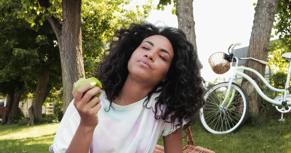 African american woman with closed eyes eating fresh apple near bicycle in park — Stock Photo