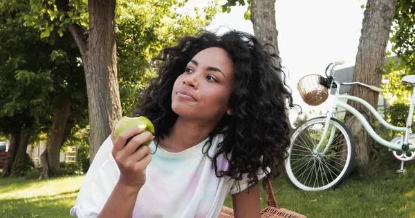 Encaracolado afro-americano mulher comendo maçã fresca perto de bicicleta no parque — Fotografia de Stock