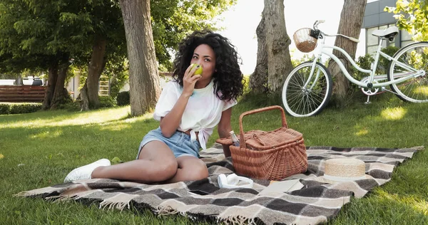 Африканская американка ест яблоко возле плетеной корзины и сидит на одеяле для пикника в парке — стоковое фото