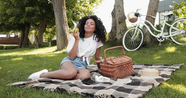 Кудрявая африканская американка ест яблоко возле плетеной корзины и сидит на одеяле для пикника в парке — стоковое фото