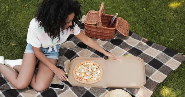 Vista de alto ângulo da mulher americana africana olhando para pizza durante piquenique no parque — Fotografia de Stock
