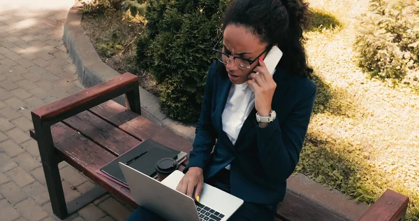 Displeased african american businesswoman talking on smartphone while sitting on bench with laptop — Stock Photo