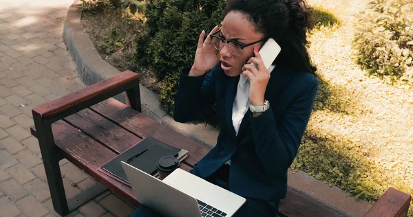 Недовольная американская деловая женщина из Африки разговаривает на смартфоне, сидя на скамейке с ноутбуком — стоковое фото