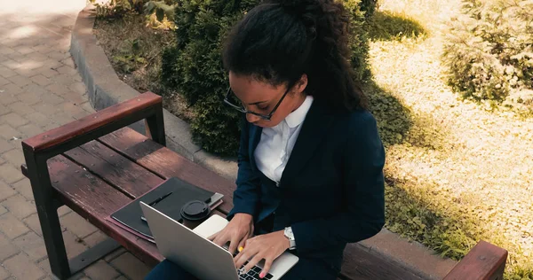 Вид зверху кучерявий афроамериканський бізнес-леді, що друкує на ноутбуці, сидячи на лавці — стокове фото