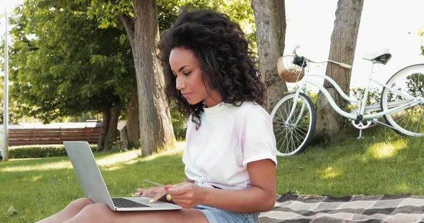 Encaracolado jovem afro-americana segurando cartão de crédito e usando laptop no parque — Fotografia de Stock