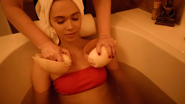 Professionelle Therapeutin massiert junge Frau mit Muscheln in Badewanne mit Wasser — Stockfoto