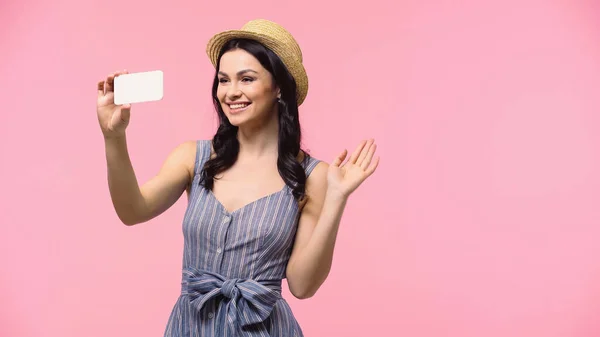 Улыбающаяся женщина в шляпе с видеозвонком на смартфоне изолирована на розовый — стоковое фото