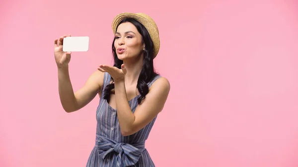 Mujer joven en sombrero de sol soplando aire beso en el teléfono inteligente aislado en rosa - foto de stock