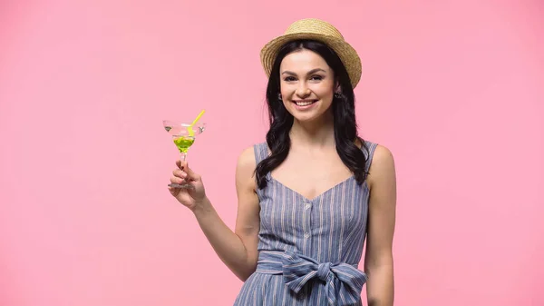 Mujer positiva en sombrero de sol sosteniendo cóctel y mirando a la cámara aislada en rosa - foto de stock