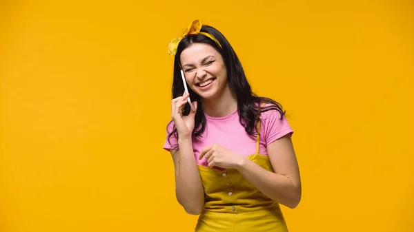 Mujer excitada con los ojos cerrados riendo mientras habla en el teléfono inteligente aislado en amarillo - foto de stock