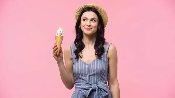 Femme rêveuse en chapeau de paille tenant la crème glacée dans le cône isolé sur rose — Photo de stock