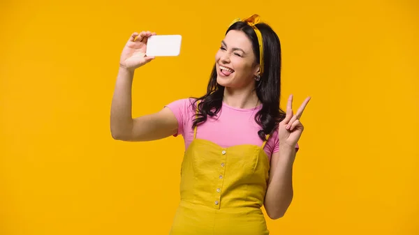 Mujer sacando la lengua y mostrando signo de paz mientras toma selfie aislado en amarillo - foto de stock